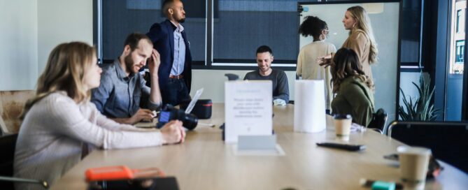 A professional business team collaborating in a modern conference room, featuring a group discussion around a wooden boardroom table with large windows in the background.